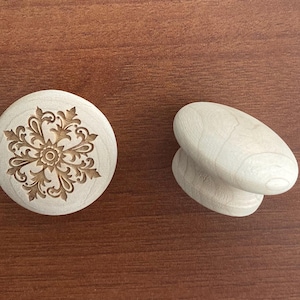 Peut inclure: Deux boutons de meuble en bois. L'un est rond avec un motif floral sculpté. L'autre est une paire de boutons de forme ovale. Les deux sont en bois de couleur claire, posés sur une surface en bois brun.