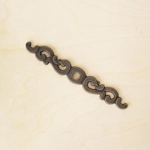 May include: An antique bronze-colored decorative furniture pull with an ornate, symmetrical design. The pull features a central circular element and scrolling details on either side, set against a light wood background.