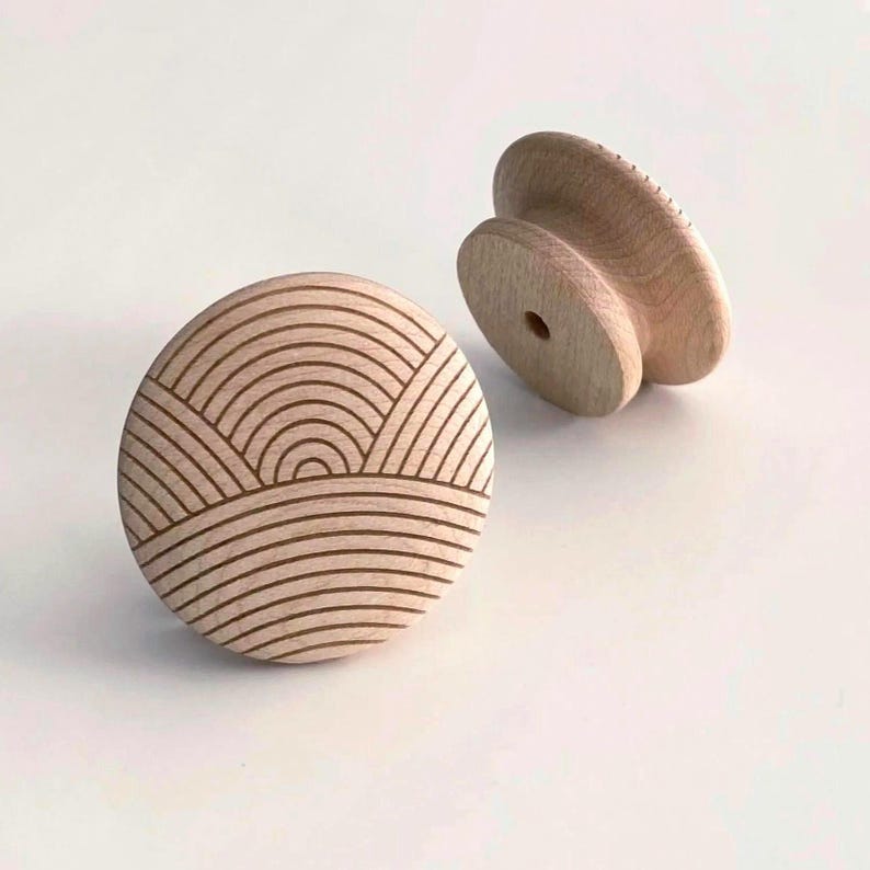 May include: Two wooden cabinet knobs. One knob is round with a carved design of curved lines resembling a landscape. The other knob is a mushroom shape with a flat top and a hole for mounting. Both knobs are light brown.