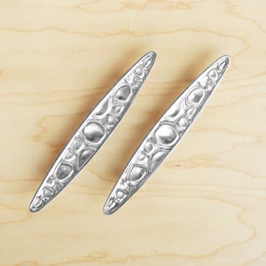May include: Two silver, decorative cabinet door pulls with a textured, raised design. The pulls are long and narrow, with a rounded end.