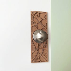 Könnte beinhalten: Dekorative Türplatte mit einem Türknauf aus Edelstahl. Die Platte besteht aus braunem Holz und weist ein ausgeschnittenes geometrisches Design auf. Der Türknauf ist rund und glänzend und steht im Kontrast zur matten Oberfläche des Holzes.