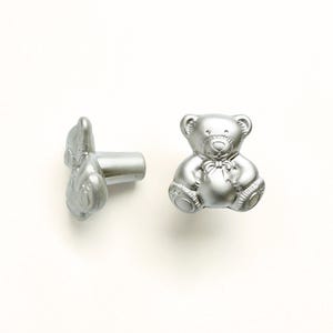 May include: Two silver teddy bear cabinet knobs. One knob is upright, and the other is on its side. The knobs have a detailed design, including a bow tie and paw details. These knobs are ideal for adding a whimsical touch to furniture.