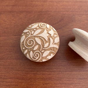 Può includere: Un bottone rotondo in legno con un design floreale dettagliato. Il bottone presenta foglie intagliate e motivi a spirale di colore marrone chiaro. Un pezzo di legno separato e non rifinito è visibile a destra.