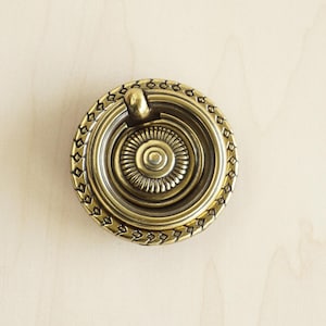 May include: A gold-toned ring pull cabinet knob with a decorative, ornate design around the edge.