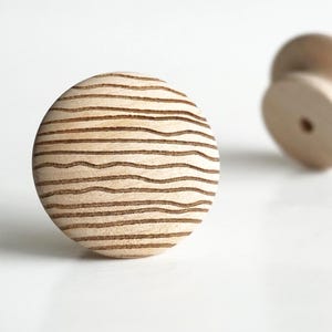 Puede incluir: Un pomo de armario redondo de madera clara con líneas horizontales grabadas. El pomo está sobre un fondo blanco, con un segundo pomo ligeramente desenfocado en el fondo.