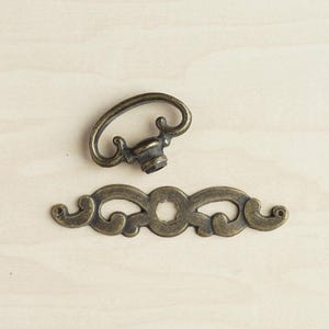 May include: An antique brass-colored metal drawer pull consisting of two separate pieces: a curved, oval-shaped handle with a central mounting point and a decorative, elongated backplate featuring symmetrical scrollwork and a central circular opening.
