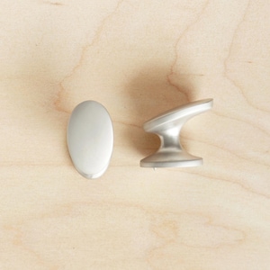 Op de afbeelding: Twee kastknoppen van geborsteld nikkel. Eén is ovaal, de andere heeft een gebogen handgreepontwerp. Ze staan tegen een lichte houtnerf achtergrond. Ideaal voor keuken- of badkamerkast upgrades.