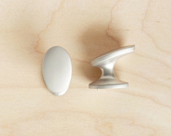 Puede incluir: Dos pomos de armario de níquel cepillado. Uno es ovalado, el otro tiene un diseño de asa curvada. Se colocan sobre un fondo claro de vetas de madera. Ideales para renovaciones de armarios de cocina o baño.