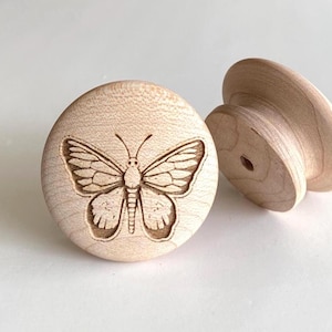 Könnte beinhalten: Zwei unfertige Holzschrankknöpfe. Ein Knopf zeigt ein detailliertes, graviertes Schmetterlingsdesign. Der andere Knopf hat eine einfache, abgerundete Form. Beide Knöpfe sind aus hellem Holz und vor weißem Hintergrund.
