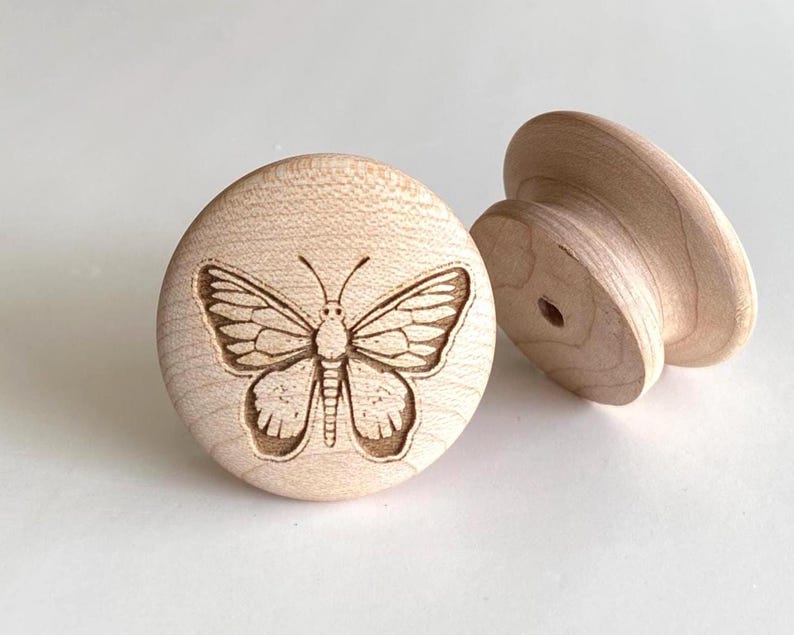 May include: Two unfinished wooden cabinet knobs. One knob features a detailed engraved butterfly design. The other knob is a simple, rounded shape. Both knobs are made of light-coloured wood and are set against a white background.