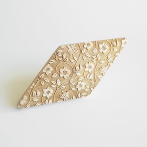 May include: A tan-coloured, diamond-shaped decorative object with a floral pattern. The design features white flowers and vines carved into the surface. The object is set against a white background.