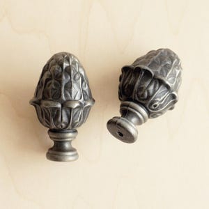 Könnte beinhalten: Zwei verzierte Metall-Abschlüsse in dunkelgrauer Ausführung. Die Abschlüsse haben ein detailliertes, florales Design und eine glatte, abgerundete Oberseite.