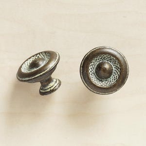 Peut inclure: Deux boutons de cabinet de style antique avec un design circulaire. Les boutons sont en métal et ont une finition vieillie avec une patine blanche.