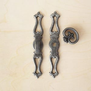 Peut inclure: Ensemble de trois pièces de quincaillerie en métal de style antique. Comprend deux longues poignées ornées et un bouton décoratif. La quincaillerie a une finition gris argenté vieilli, disposée sur une surface en bois clair.