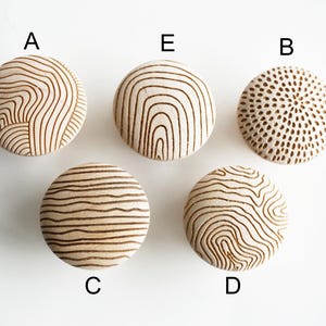 Op de afbeelding: Vijf ronde, lichtgekleurde houten knoppen met verschillende lasergegraveerde patronen. Elke knop is gelabeld met een letter: A, B, C, D en E. De patronen omvatten diverse topografische lijnen, concentrische cirkels en gestippelde texturen.