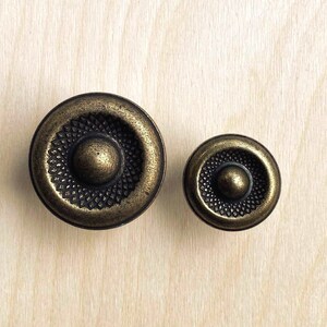 May include: Two round cabinet knobs with an antique brass finish. Each knob features a central dome surrounded by a textured, cross-hatched pattern within a smooth outer rim. The knobs are circular in shape and display a weathered, metallic appearance.