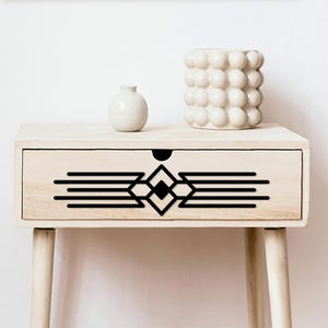 May include: A light-colored wooden nightstand with a drawer featuring a black geometric design. Two white ceramic decorative pieces sit on top. The nightstand has four tapered legs, offering a modern aesthetic.