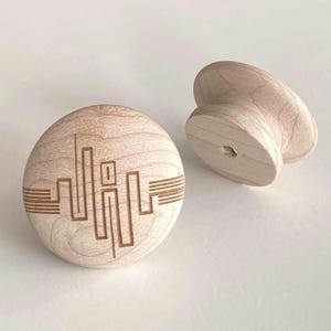 Op de afbeelding: Twee onafgewerkte houten kastknoppen. Eén is rond met een stadsgezichtontwerp in het oppervlak geëtst. De andere knop is een gebogen, hartvormig ontwerp. Beide knoppen zijn gemaakt van lichtgekleurd hout.