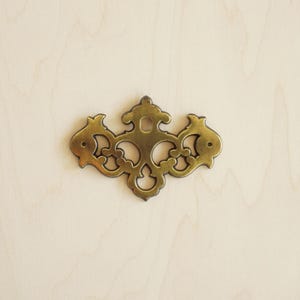 May include: A gold-toned metal drawer pull with a decorative, ornate design. The pull has a central oval opening and intricate scrollwork.