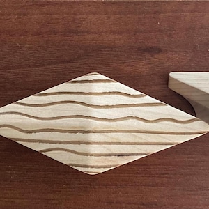 以下が含まれることがあります： 2つの木製キャビネットプル。1つは波状の線がエッチングされたひし形で、もう1つは幾何学的な形状です。どちらも明るい色の木材で、濃い色の木材の表面に置かれています。