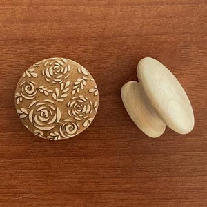 Könnte beinhalten: Ein runder Holzknopf mit floralem Design und zwei ovale Holzknöpfe. Der runde Knopf hat einen hellbraunen Hintergrund mit einem geschnitzten Blumenmuster. Die ovalen Knöpfe sind hell und naturfarben.