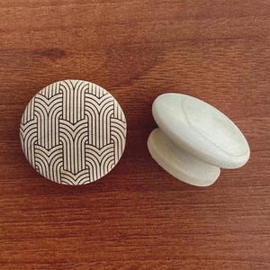 Könnte beinhalten: Zwei dekorative Holzobjekte auf einer braunen Oberfläche. Eines ist ein runder Knopf mit einem schwarz-weißen geometrischen Muster. Das andere sind zwei gestapelte, oval geformte Holzstücke. Die Objekte sind hell.