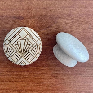 Könnte beinhalten: Ein runder Holzknopf mit Art-Deco-Design und zwei ovale Holzknöpfe. Der runde Knopf hat ein geometrisches Muster, das in seine Oberfläche geschnitzt ist. Die Knöpfe sind hell und liegen auf einer braunen Holzoberfläche.