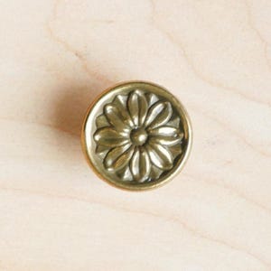 Może przedstawiać: Złoty metalowy gałka z kwiatowym wzorem. Gałka ma okrągły kształt i wypukły, szczegółowy wzór kwiatu.