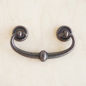 May include: A dark brown metal drawer pull with a rounded, decorative center. The pull has a loop shape and is attached to a round base.