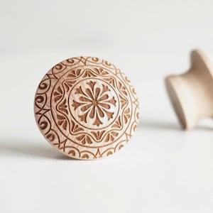 Puede incluir: Tirador de armario de madera marrón claro con un intrincado diseño floral y geométrico tallado. El tirador circular presenta un motivo floral central rodeado de patrones detallados. Otro tirador está ligeramente desenfocado en el fondo.