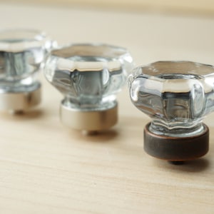 Könnte beinhalten: Drei klare Glas-Schrankknöpfe mit verschiedenen farbigen Metallsockeln. Die Knöpfe sind rund mit einem facettierten Design. Die Sockel sind silber, silber und bronzefarben.