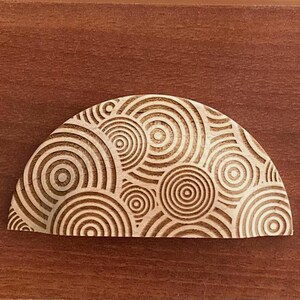 Peut inclure: Un objet en bois clair, semi-circulaire, avec un motif de cercles concentriques répétés. Le motif est sculpté dans le bois, créant une surface texturée. L'objet est présenté sur une surface en bois brun foncé.