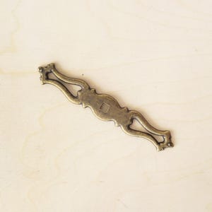 Pode incluir: Uma puxador de gaveta de metal dourado com um design ornamentado. O puxador tem um furo quadrado no centro para montagem.