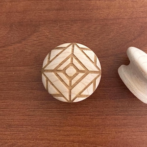 Peut inclure: Un objet rond en bois clair avec un motif géométrique gravé. Le motif présente un losange central entouré de lignes obliques. Un autre objet en bois incurvé est également visible.