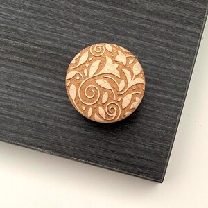 May include: A round, wooden decorative knob with an engraved floral design. The knob is light brown and features a pattern of leaves and swirls. It rests on a dark gray surface.