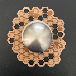 May include: A decorative wooden coaster with a honeycomb pattern and a stainless steel, spherical object in the center. A detailed illustration of a bee is on the right side of the coaster. The coaster is brown and the background is black.