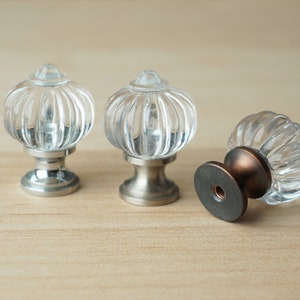 May include: Three clear glass cabinet knobs with a ribbed design. Two knobs have a silver finish and one has a bronze finish.