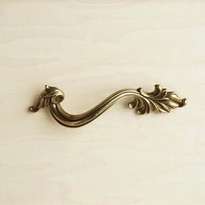 May include: A brass-toned metal drawer pull featuring an S-shaped curved body with ornate, leaf-like detailing at both ends. The hardware has a smooth, polished finish with darker recessed areas that highlight the intricate, sculpted metal design.