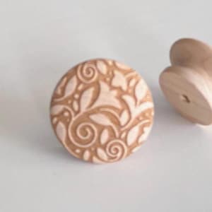 May include: A light brown wooden cabinet knob with a floral design. The knob is round with a carved pattern of leaves and swirls. A second, unfinished knob is visible in the background.