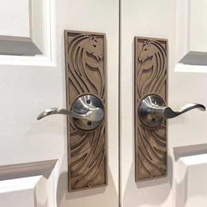 May include: Wooden door handle plates featuring a horse head design. The plates are light brown and mounted on a white door. The handles are silver-coloured and curved.