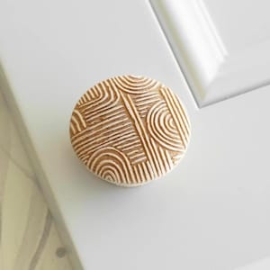 Peut inclure: Un bouton de meuble rond, brun clair, avec un motif géométrique sculpté de lignes et d'arcs. Le bouton est posé sur une porte d'armoire blanche, offrant un contraste neutre. Cette quincaillerie décorative ajoute une touche de design moderne.