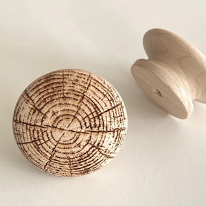 Puede incluir: Dos tiradores de armario de madera. Uno es redondo con un patrón detallado de vetas de madera. El otro es un tirador curvo, liso y de color claro. Ambos son de madera sin terminar y sobre un fondo blanco.