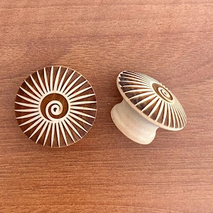May include: Two decorative wooden cabinet knobs with a spiral and sunburst design. The knobs are brown with white accents and are set against a brown wooden surface. The knobs are approximately 2 inches in diameter.