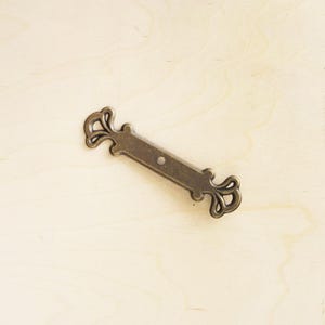 May include: A decorative metal drawer pull with a simple, elegant design. The pull is made of a dark, aged metal and has a slightly ornate, floral pattern on each end.