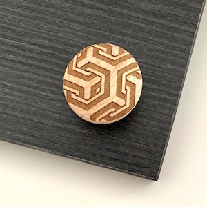 Könnte beinhalten: Ein runder Holzknopf mit geometrischem Design. Der Knopf ist hellbraun mit einem geschnitzten Muster aus ineinandergreifenden Formen. Er liegt auf einer dunkelgrauen Oberfläche.