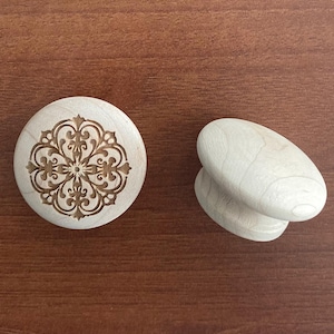 Könnte beinhalten: Zwei hölzerne Schrankknöpfe. Einer ist rund mit einem geschnitzten Blumendesign in einem dunkleren Holzton. Der andere Knopf ist oval geformt mit einer glatten, hellen Holzoberfläche. Beide sind auf einer braunen Holzoberfläche platziert.