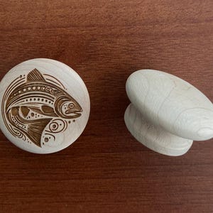 May include: Two wooden cabinet knobs. One is round with a detailed etched design of a fish. The other knob is oval-shaped with a smooth, unfinished surface. Both are light-coloured wood.