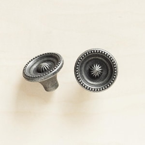 Op de afbeelding: Twee zilverkleurige metalen kastknoppen met een decoratief, verhoogd stervormig ontwerp in het midden. De knoppen hebben een parelrand.