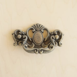 Peut inclure: Une poignée de tiroir décorative, ornée, en métal avec un centre circulaire surélevé. La poignée est finie dans une couleur laiton brossé antique.