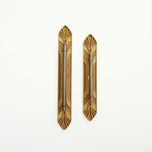 May include: Two gold-toned door handles with an Art Deco design. The handles are rectangular with a decorative pattern and a vertical bar in the centre.